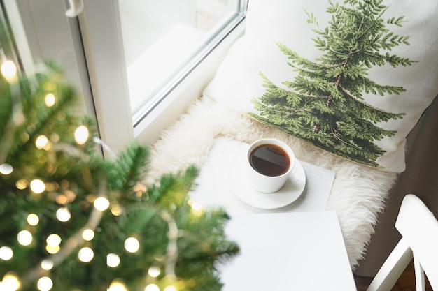 Café de invierno en el alféizar de la ventana con árbol de Navidad y almohada para una lectura acogedora