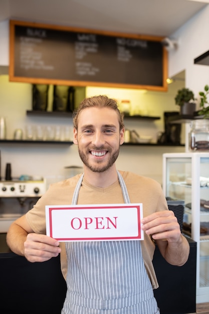 Cafe ist geöffnet. Glücklicher optimistischer junger erwachsener Mann in der Schürze, die lehnendes Barzählerhalteschild offen im Café steht