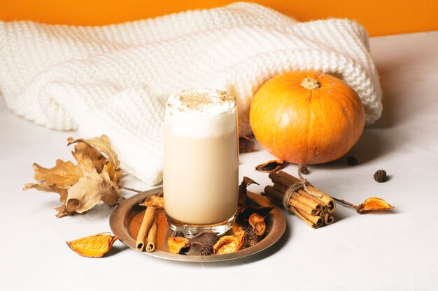 Un café con leche de calabaza y especias se encuentra junto a una bufanda de punto, canela, hojas secas y calabaza.