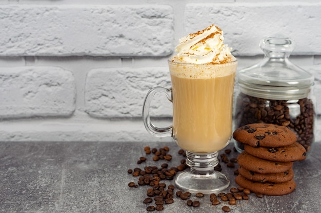 Café con leche de caramelo en un vaso alto con crema batida y galleta de chocolate. Granos de café en la superficie de la pared de ladrillo gris y blanco con espacio de copia.