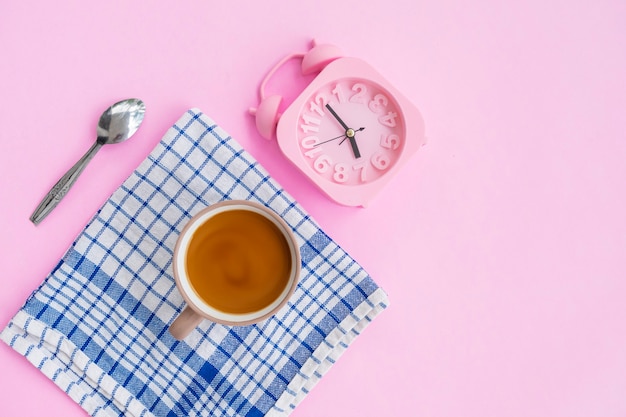 Café con leche, cuchara, despertador y paño azul aislado sobre fondo rosa, idea de concepto mínimo.