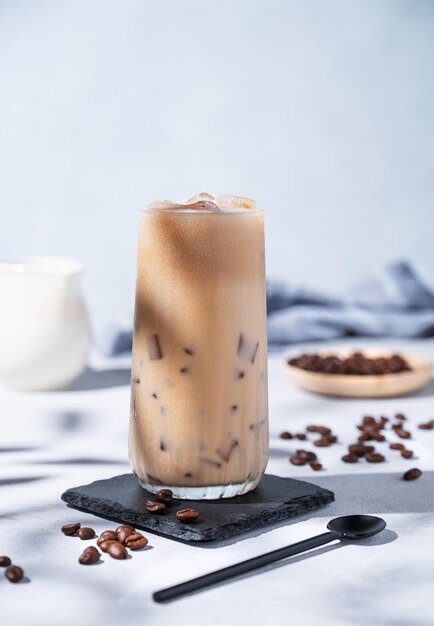 Café con leche helado en un vaso alto con leche sobre un fondo azul con frijoles de café, jarro de leche y sombras matutinas Concepto de refresco de verano