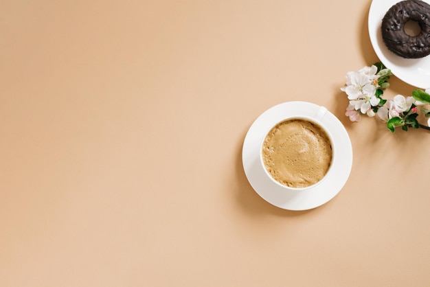 Café con leche en una taza de porcelana blanca y donuts de chocolate con flores de manzana blanca sobre un fondo beige El concepto de desayuno matutino o descanso para el café