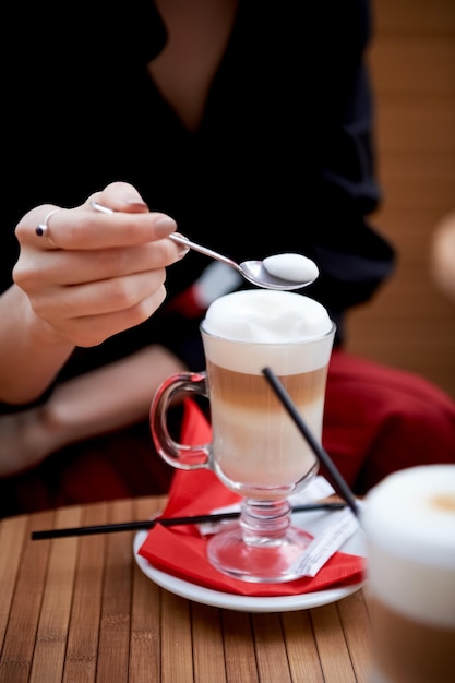 Café con leche en taza de vidrio con mango de espuma alta De pie sobre una servilleta roja Manos con una cuchara