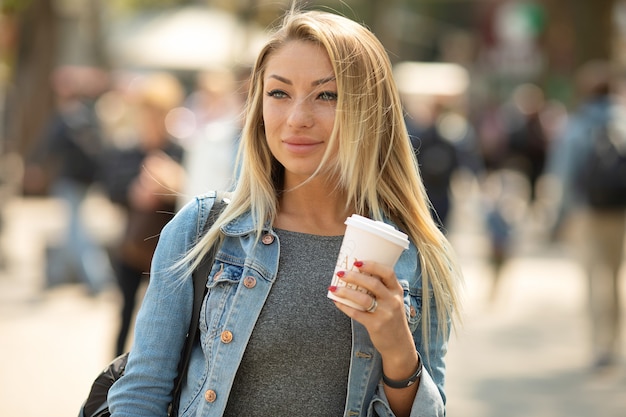 Café para llevar. Hermosa mujer joven sosteniendo la taza de café y sonriendo mientras camina por la calle.