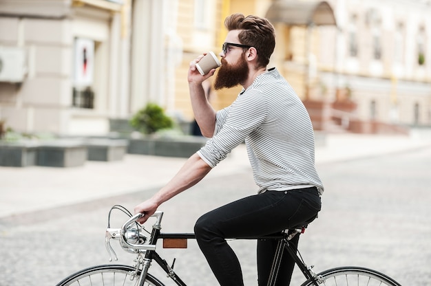 Café para llevar. Vista lateral del joven barbudo tomando café mientras está sentado en su bicicleta al aire libre