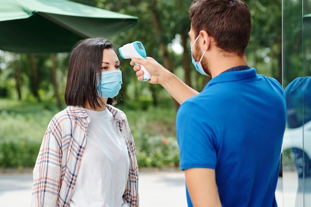 Café-Manager, der die Körpertemperatur einer Kundin in einer medizinischen Maske mit einem berührungslosen Infrarot-Thermometer überprüft