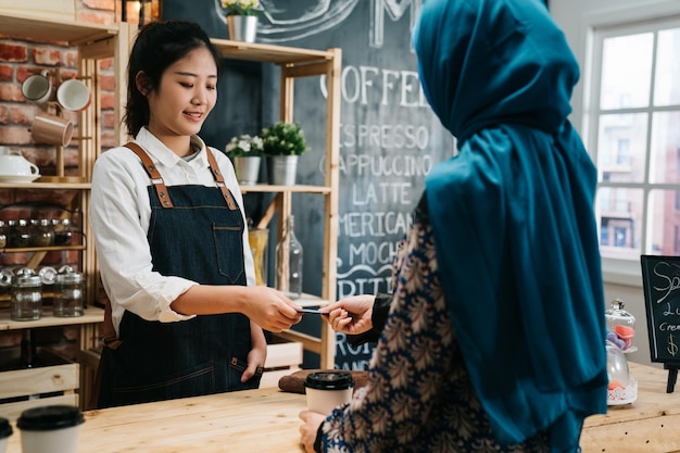 El café moderno pone en marcha el concepto de micropago para pequeñas empresas. Una camarera interactuando con un cliente islámico usando una terminal bancaria para procesar y adquirir pagos con tarjeta de crédito en una cafetería en un día soleado.