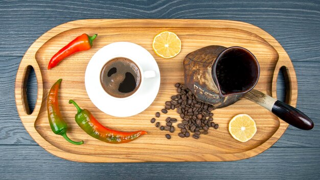 Café negro caliente recién hecho en una taza blanca sobre una tabla de madera junto a un pavo de cerámica con granos de café. pimiento picante y limón