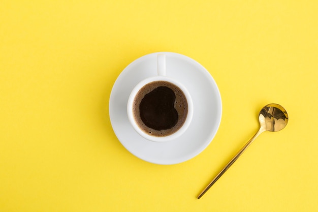 Café negro en la taza blanca y cuchara dorada en el centro de la mesa amarilla. Copie el espacio. Vista superior.