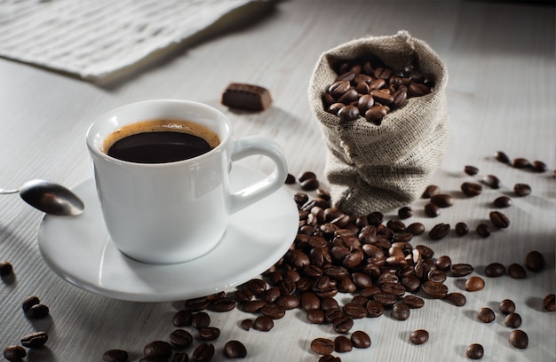 Café negro en una taza blanca en un platillo. Granos de café en la bolsa y esparcidos sobre la mesa.