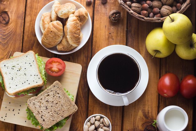 Café negro en una taza blanca sobre una mesa de madera. Desayuno en el pueblo. bocadillos con verduras y hierbas, leche en lechero, frutos secos y frutas con dulces.