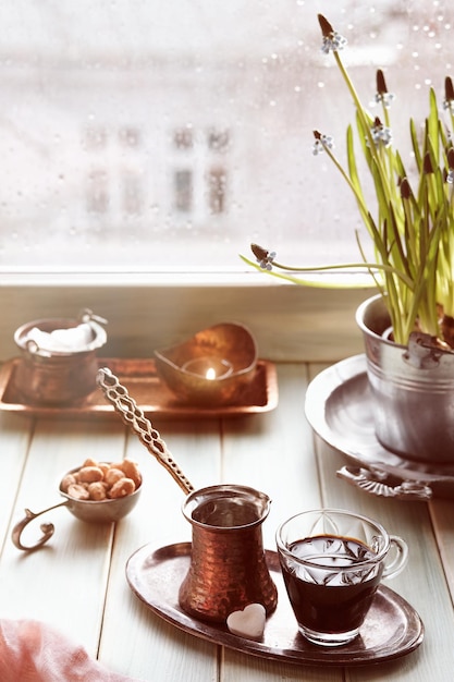 Café oriental na cafeteira de cobre turca tradicional com flores no parapeito da janela. Peitoril da janela de madeira com vaso de flores jacinto. Dia frio e chuvoso na primavera. Cena aconchegante, conceito de higiene.