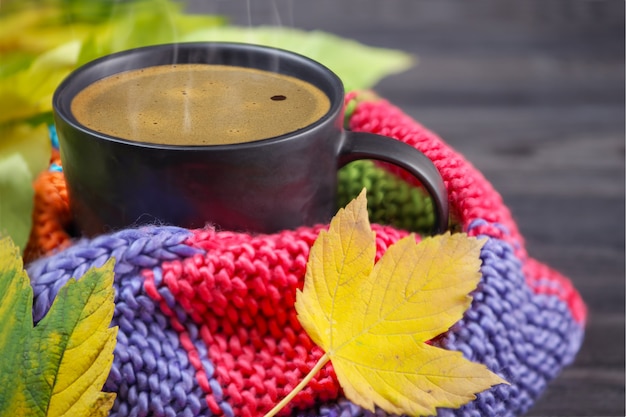 Café preto em uma caneca preta embrulhada em um cachecol quente, colorido, de malha e folhas de outono