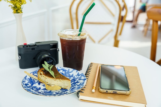 Café preto gelado americano na xícara caneca cheesecake de rosto queimado colocado e câmera smartphone no notebook na mesa de madeira na vista superior como café da manhã em uma cafeteria no conceito de trabalho de negócios de cafedura