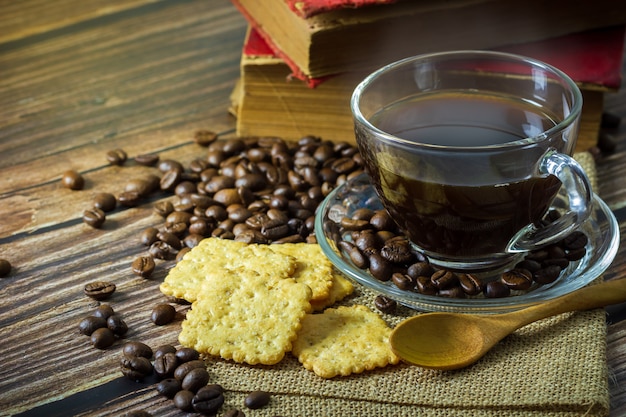 Café preto no copo e nos feijões de café de vidro claros com biscoitos e livro velho na tabela de madeira.