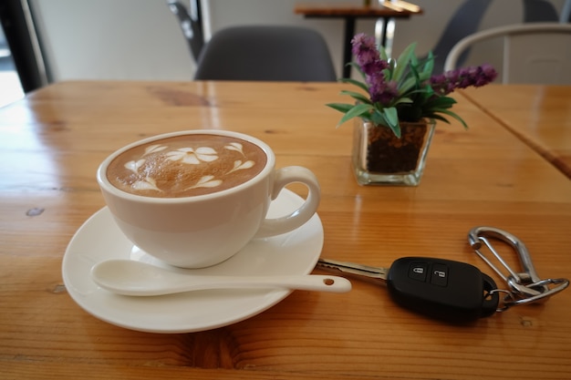 Café quente, cappuccino em uma xícara de café branco em uma mesa de madeira de pinho E há uma chave de carro ao lado dela