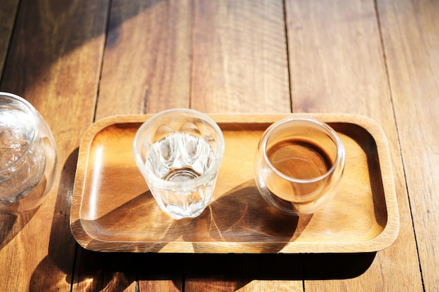 Café quente e copo de gelo na bandeja de madeira sob a luz solar