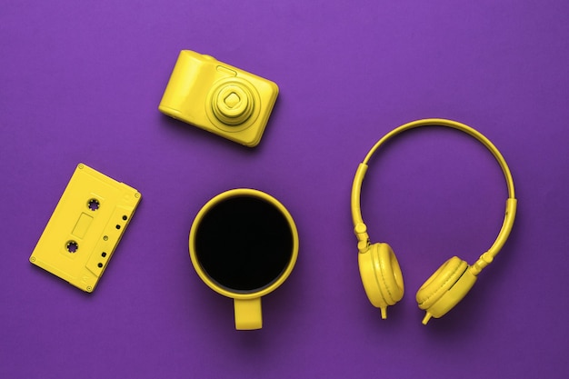 Café quente em uma caneca amarela, fones de ouvido, uma fita cassete e uma câmera em um fundo roxo. Tendência de cores.
