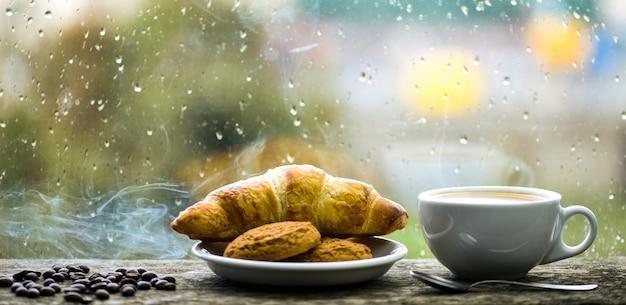 Café recién hecho en taza blanca o taza en el alféizar de la ventana Bebida de café con postre de croissant Disfrutando del café en un día lluvioso Hora del café en un día lluvioso Ventana de vidrio mojado y taza de bebida de cafeína caliente