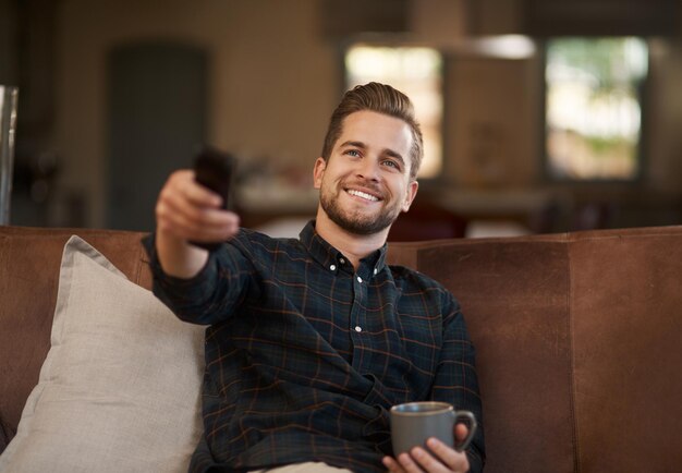 Foto café relajante y un hombre viendo la televisión en un sofá en la sala de estar de su casa con un control remoto para transmitir té sonriente y un joven feliz usando un servicio de suscripción en su televisión