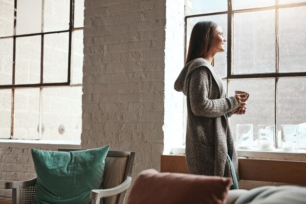 Café relaxante e mulher olhando pela janela de sua casa pensando e sonhando acordada em uma manhã tranquila Chá de devaneio e menina olhando contemplando e descansando em sua sala de estar no fim de semana