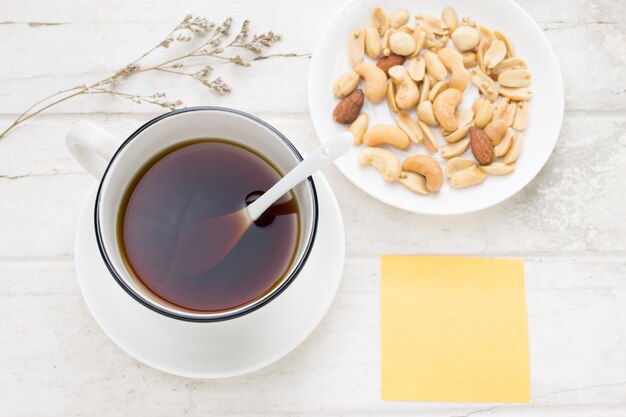 Café sólo en la taza blanca y nueces mezcladas con la cuchara blanca, la flor secada y la nota pegajosa en la tabla blanca. Vista superior.