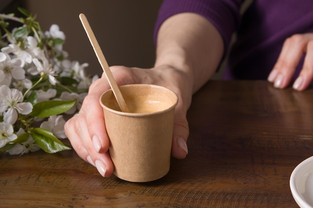 Café en una taza de cartón para llevar con un agitador en una mano femenina sobre una mesa con flores