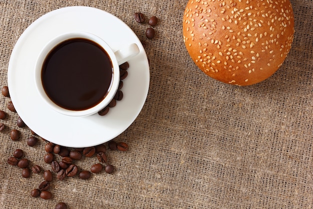 Café en una taza y plato, bollo con sésamo y grano de café sobre mantel de arpillera. Vista superior.