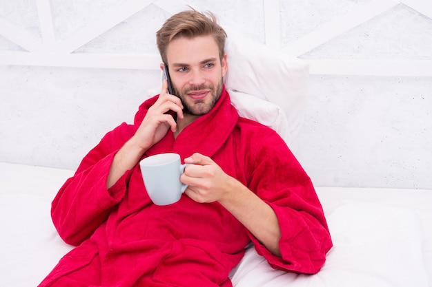 El café y la tecnología han ido de la mano Joven usando tecnología móvil en la cama Chico guapo hablando por teléfono y bebiendo café en casa Vida moderna y nueva tecnología Concepto de tecnología