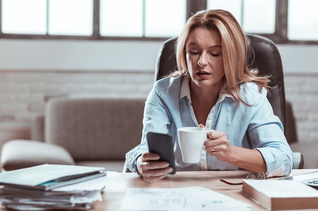 Café y teléfono. Empresaria madura de pelo rubio disfrutando de un descanso tomando café y usando el teléfono inteligente