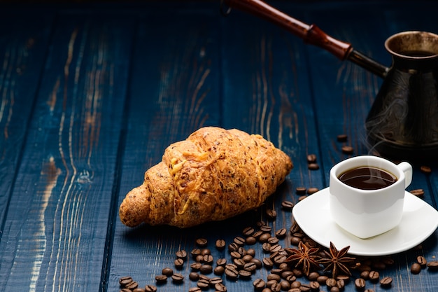 El café se vierte en una taza sobre una mesa de madera azul con granos de café y un croissant.