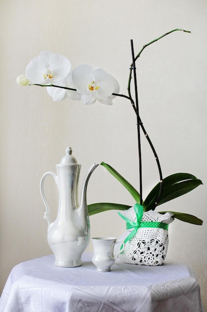 Cafeteira branca com uma flor branca na mesa com uma toalha de mesa branca