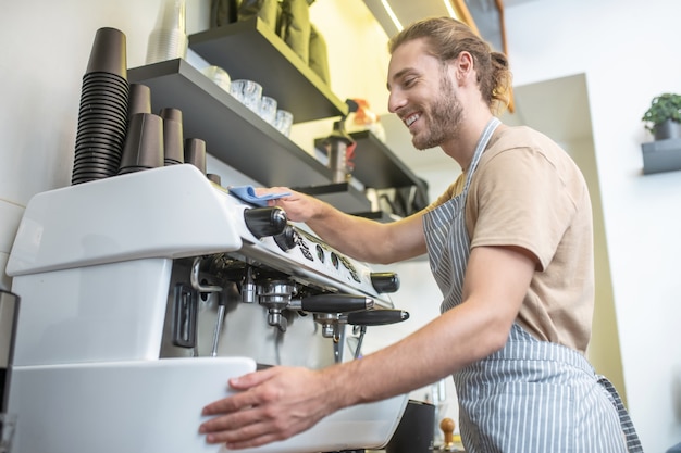 Cafetera, limpieza. Hombre alegre en un delantal limpiando cuidadosamente la máquina de café en su café