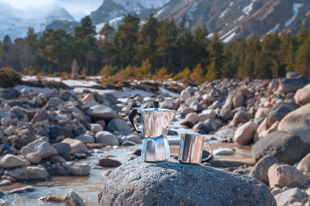 Cafetera moka y taza de metal en piedra grande contra el telón de fondo del río de montaña