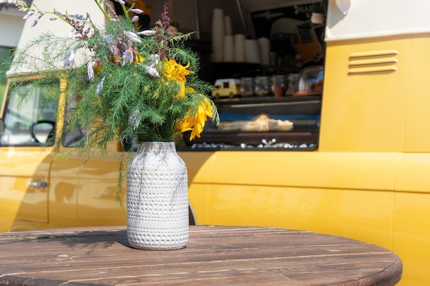 Cafetería de la calle amarilla sobre ruedas junto a la mesa con flores. Camión de comida vintage con mesa redonda vacía en un día soleado.