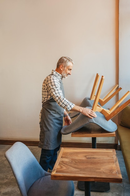 Cafetería, limpieza. Hombre de cabello gris en delantal de pie de lado a la silla de elevación de la cámara en la mesa en el café a la luz del día