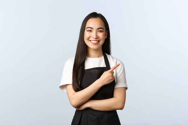 Cafetería, pequeña empresa y concepto de puesta en marcha. Chica asiática alegre en restaurante o cafetería de trabajo delantal, invitando a los consumidores a ofertas especiales. Personal femenino sonriente señalando con el dedo a la derecha.