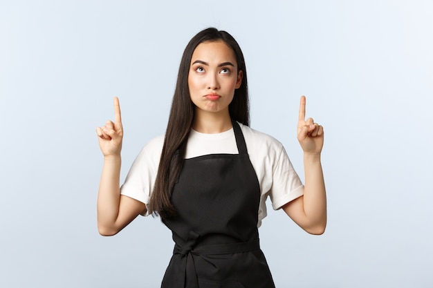 Cafetería, pequeña empresa y concepto de puesta en marcha. Decepcionado, sombrío y lindo barista de sexo femenino asiático apuntando con el dedo hacia arriba con una mueca de malestar. Chica que trabaja en restaurante en delantal, expresa disgusto o arrepentimiento.