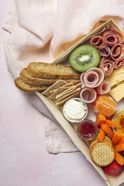 Caixa de brunch saudável para levar com presunto, morangos; Kiwi; pão; biscoitos; queijo, cenoura, tangerina, homus.