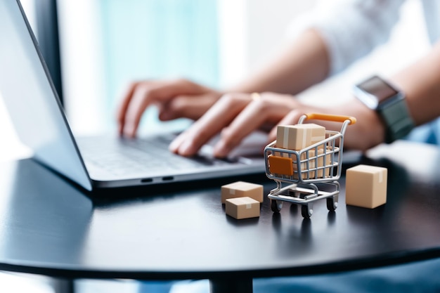 Foto caixa de encomendas no carrinho na mesa com a mão de uma jovem usando laptop para fazer compras