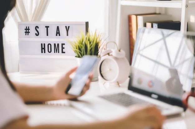 Caixa de luz com a hashtag de texto #STAYHOME brilhando na luz e turva mulher trabalhando em casa. Trabalhador de escritório em quarentena. Trabalho em casa para evitar doenças virais. Freelancer ou conceito de trabalhador remoto.