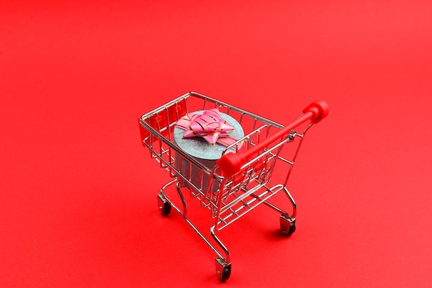 Caixa de presente azul com laço rosa em um carrinho de compras em um fundo vermelho.