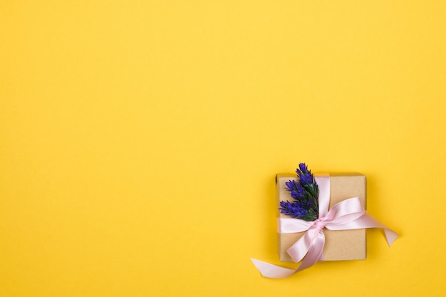 Caixa de presente de papel com um laço rosa e um galho de lavanda no canto em uma parede amarela