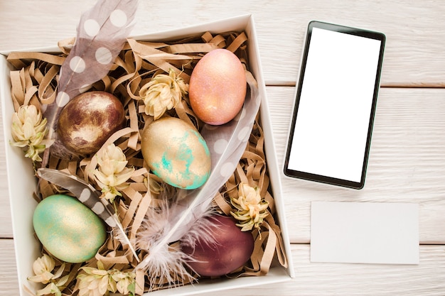 Caixa de presente de Páscoa com ovos de cor, penas e flores na mesa de madeira branca. Smartphone com tela em branco e cartão vazio com espaço livre para texto.