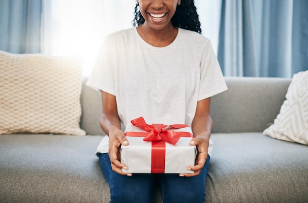 Caixa de presente e mulher em um sofá na sala de estar com um gesto de doação para celebração ou evento Papel de embrulho de laço e mulher africana com um presente de aniversário de natal ou feriados em casa