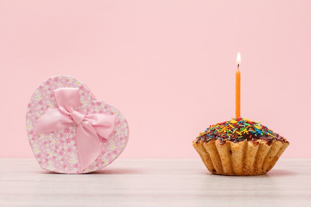 Caixa de presente em forma de coração e saboroso muffin de aniversário com esmalte de chocolate e caramelo, decorada com uma vela festiva acesa no fundo de madeira e rosa. Conceito mínimo de feliz aniversário.