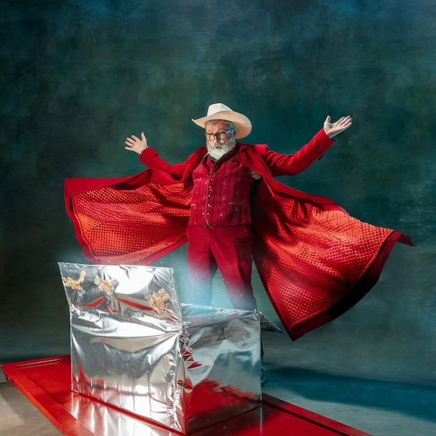 Caixa de presente mágica. Homem moderno e elegante em um terno vermelho elegante e chapéu de cowboy em fundo escuro.