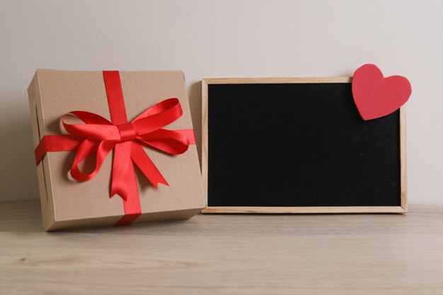 Caixa de presente marrom com fita vermelha e quadro-negro em fundo de madeira.