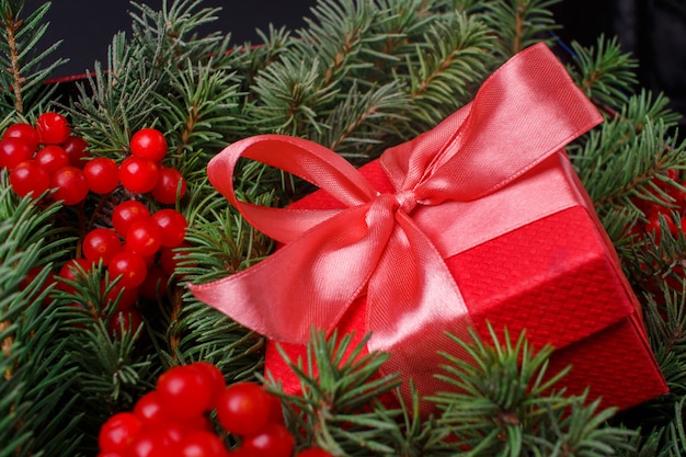 Caixa de presente vermelha com laço de cetim rosa, imersa nas agulhas de uma árvore de Natal decorada com frutas vermelhas.